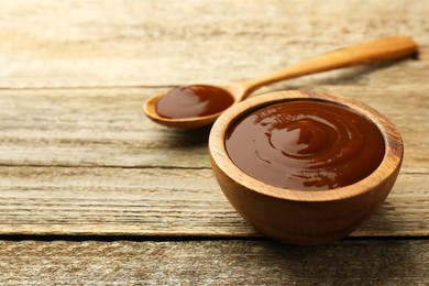 Tasty barbeque sauce in bowl and spoon on wooden table, closeup. Space for text