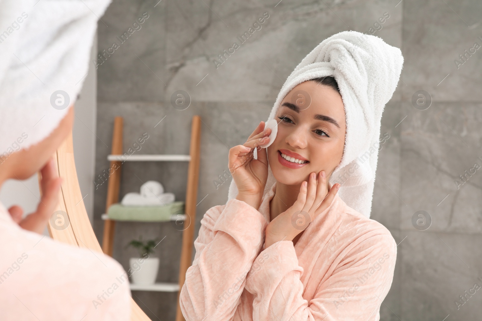 Photo of Beautiful woman in terry towel removing makeup with cotton pad near mirror indoors