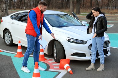 Photo of Young woman with instructor near car and fallen traffic cone outdoors. Failed driving school exam