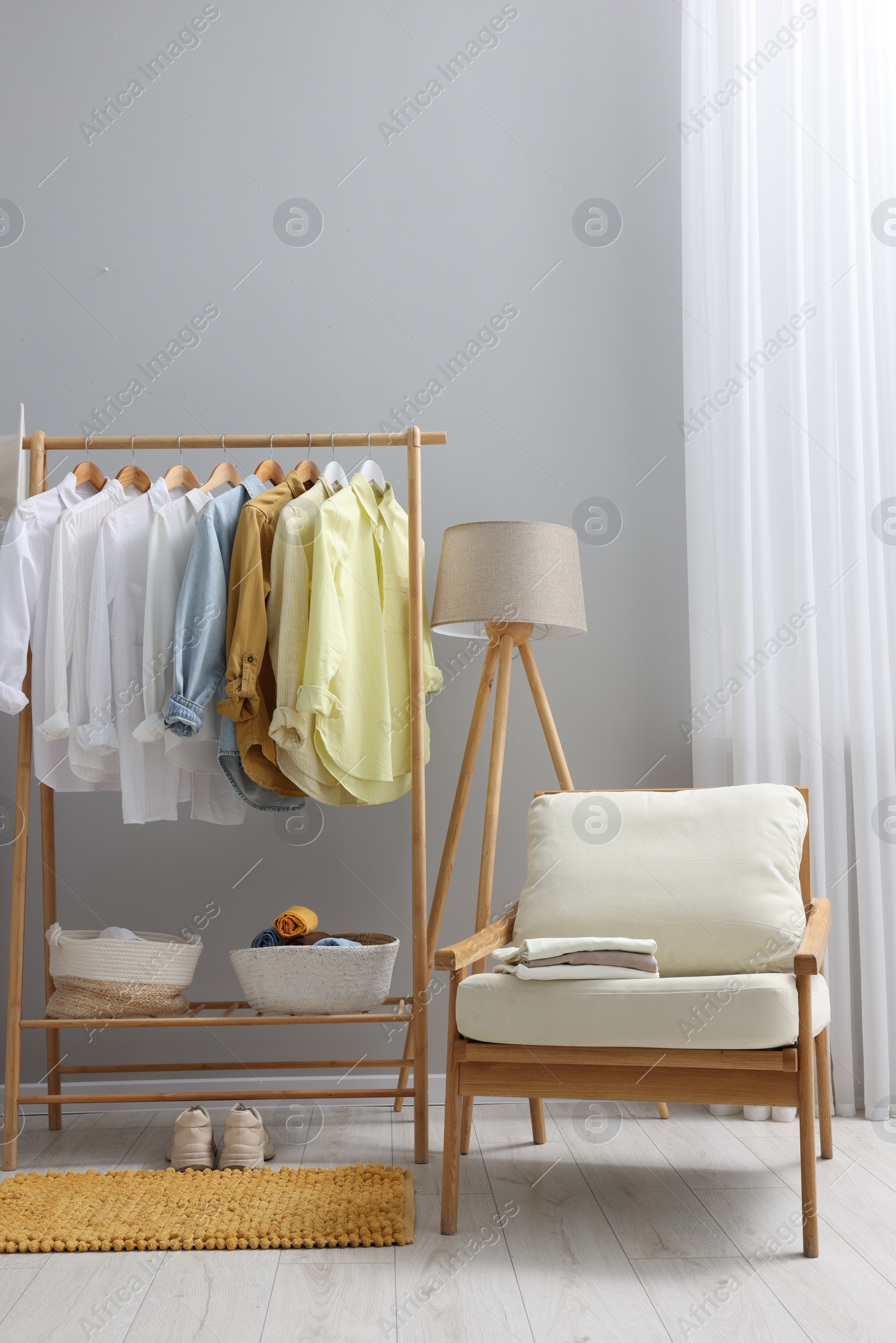 Photo of Wardrobe organization. Rack with different stylish clothes, armchair and lamp near grey wall indoors