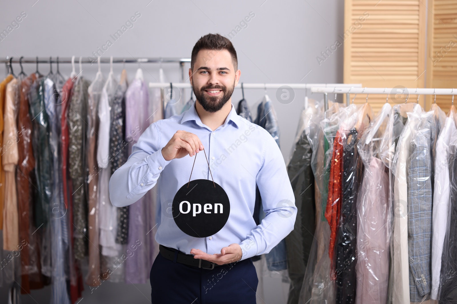 Photo of Dry-cleaning service. Happy worker holding Open sign near racks with clothes indoors