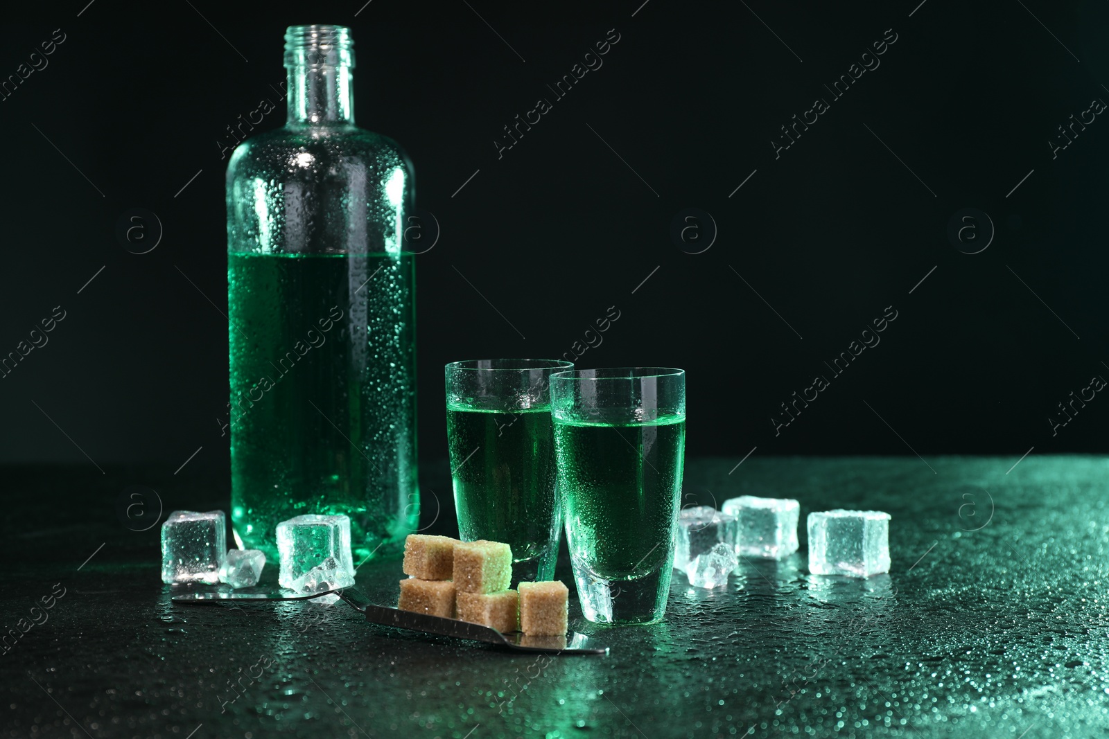 Photo of Absinthe in shot glasses, spoon, brown sugar and ice cubes on gray table against dark background, space for text. Alcoholic drink