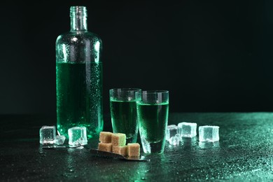 Absinthe in shot glasses, spoon, brown sugar and ice cubes on gray table against dark background, space for text. Alcoholic drink