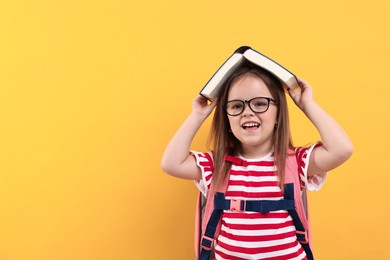 Photo of Cute little girl in glasses with open book and backpack against orange background. Space for text