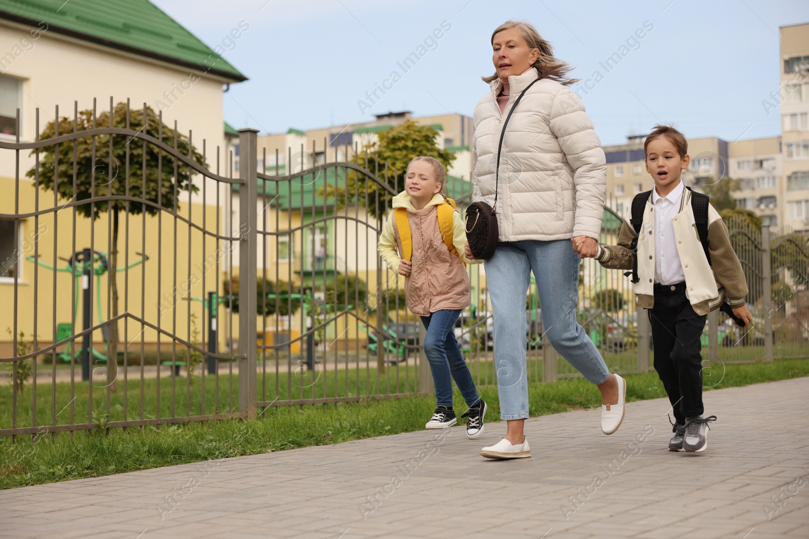 Photo of Being late for school. Senior woman and her grandchildren with backpacks running outdoors, low angle view