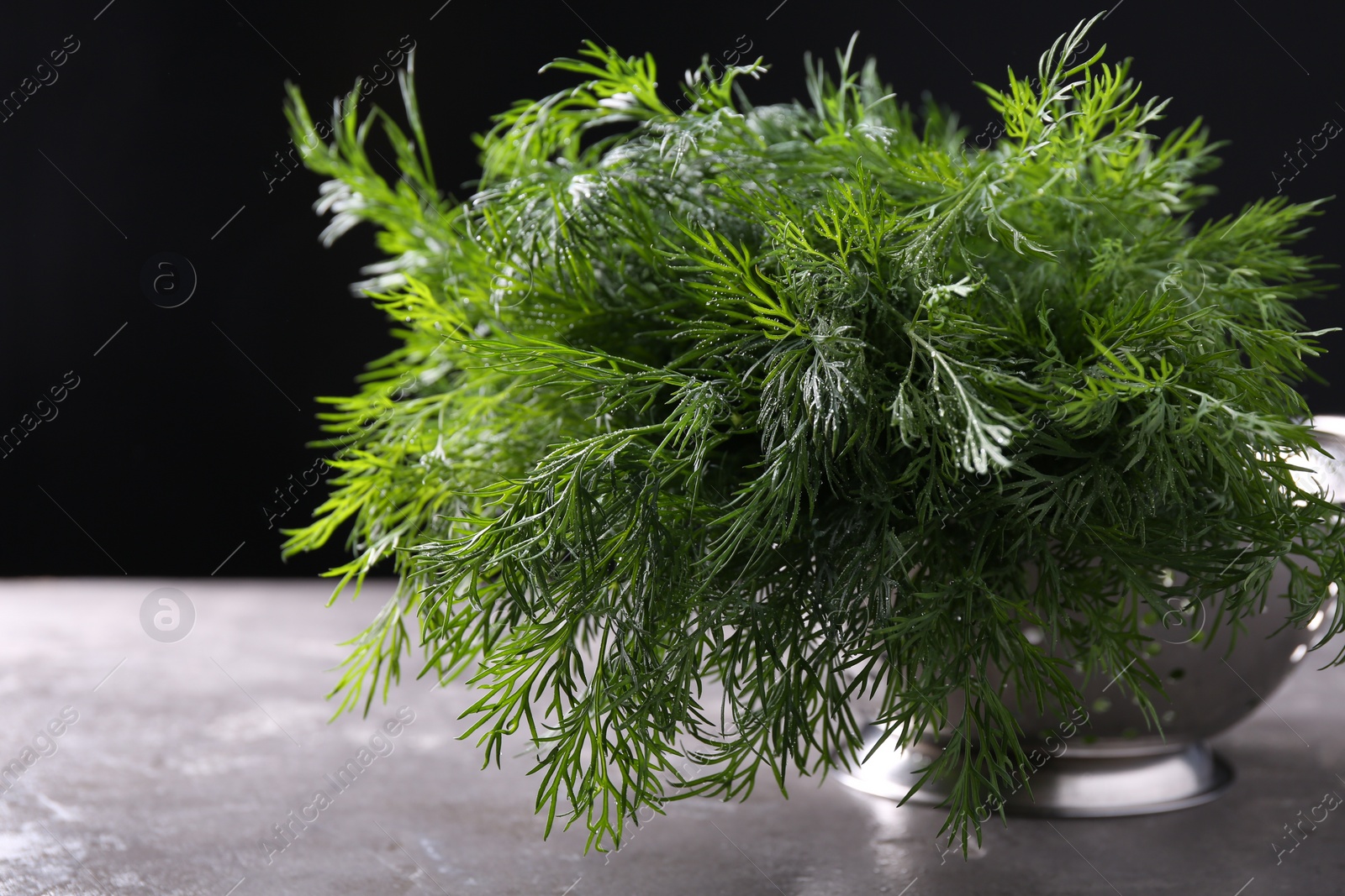 Photo of Fresh wet dill in colander on grey textured table against black background, closeup