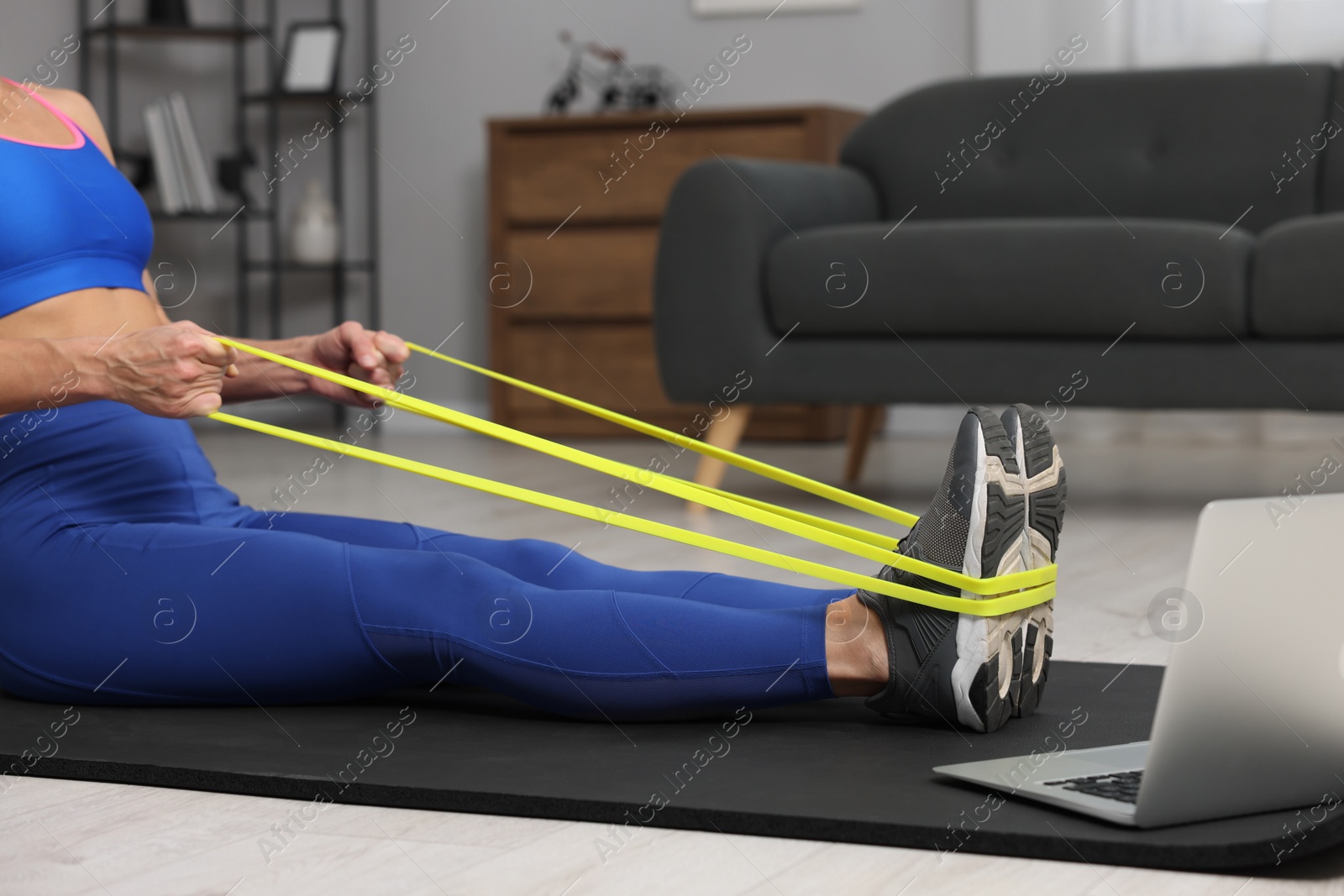 Photo of Woman doing exercise with fitness elastic band near laptop on mat at home, closeup