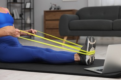 Woman doing exercise with fitness elastic band near laptop on mat at home, closeup