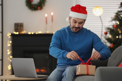 Photo of Celebrating Christmas online with exchanged by mail presents. Man in Santa hat opening gift box during video call on laptop at home