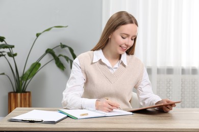 Photo of Woman taking notes while using tablet at wooden table in office