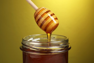 Photo of Pouring tasty honey from dipper into glass jar on golden background, closeup