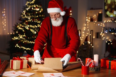 Photo of Santa Claus wrapping gift at his workplace in room decorated for Christmas