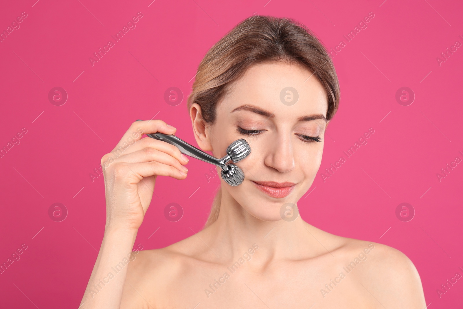 Photo of Young woman using metal face roller on pink background