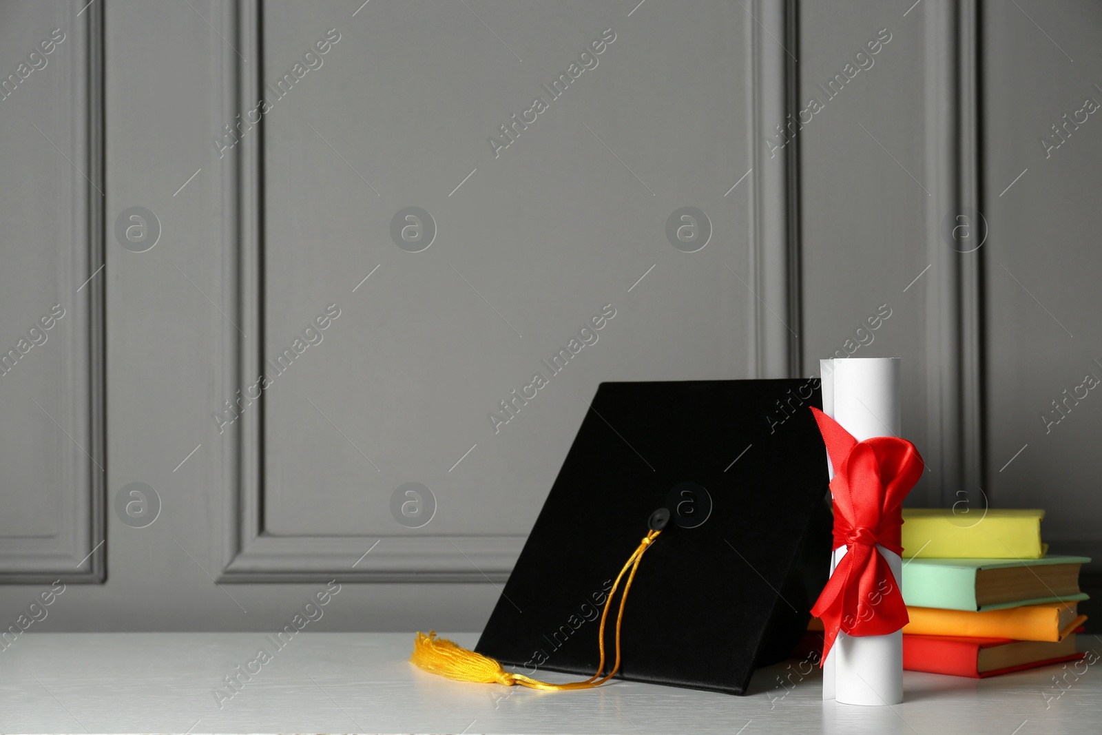 Photo of Graduation hat, books and diploma on white table near grey wall, space for text