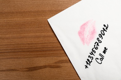 Paper napkin with phone number and lipstick mark on wooden table, top view. Space for text