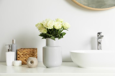 Vase with beautiful white roses and toiletries near sink in bathroom