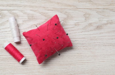 Photo of Red pincushion with sewing pins and spools of threads on light wooden table, flat lay. Space for text