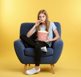 Photo of Emotional teenage girl with popcorn sitting in armchair during cinema show on color background