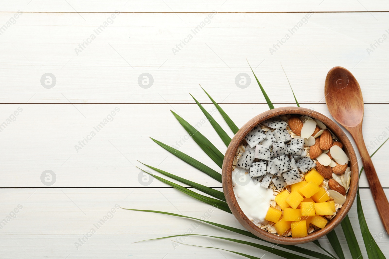 Photo of Bowl of granola with pitahaya, mango, almonds and yogurt on white wooden table, flat lay. Space for text