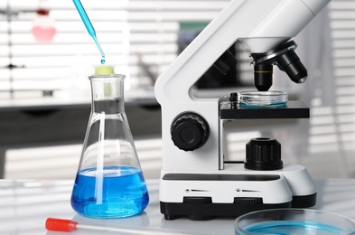 Photo of Laboratory analysis. Dripping blue liquid into flask near microscope on white table, closeup