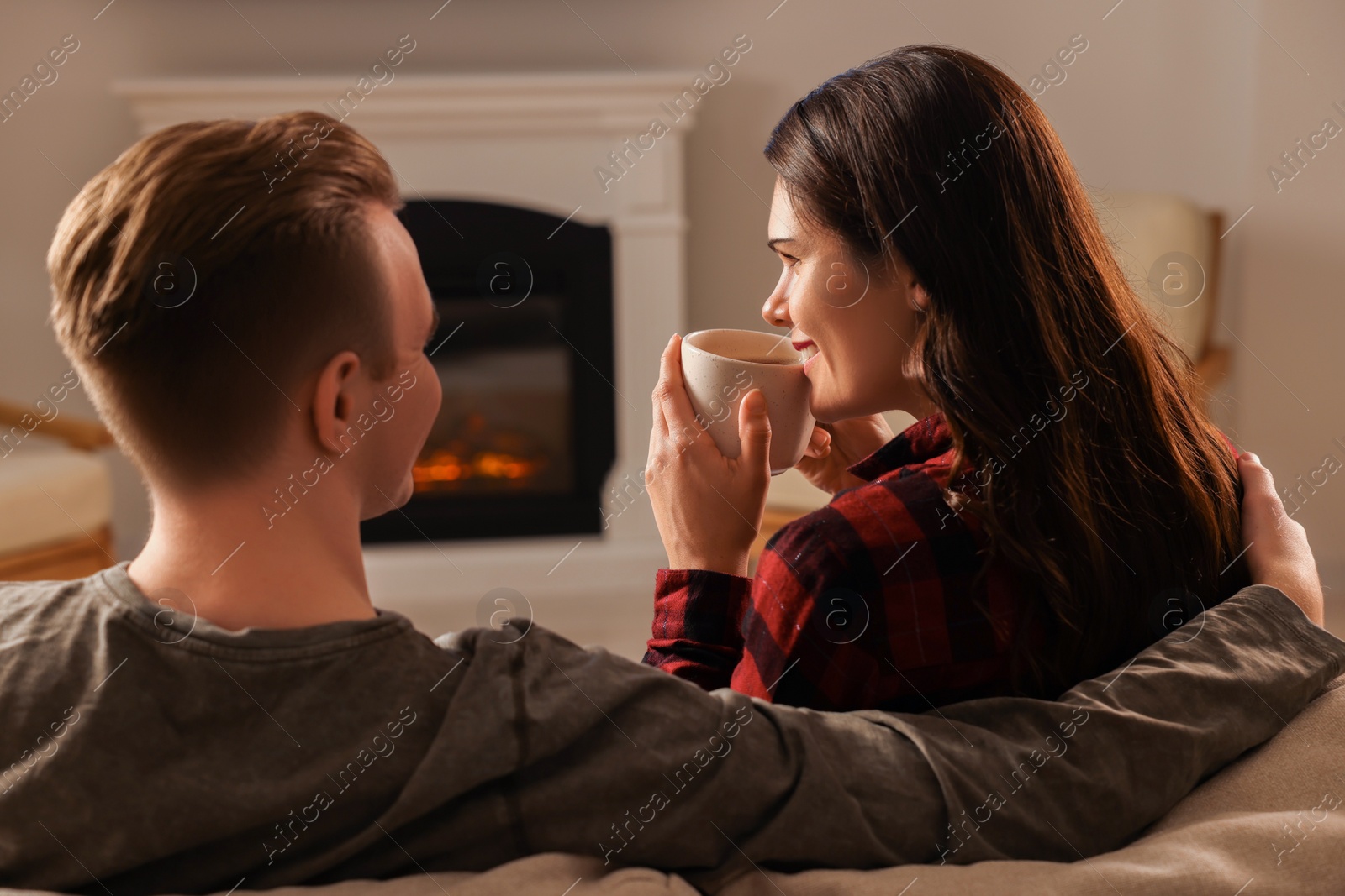 Photo of Happy lovely couple with hot drinks spending time together near fireplace at home, back view