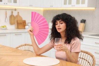 Photo of Young woman with glass of water waving pink hand fan to cool herself at table in kitchen