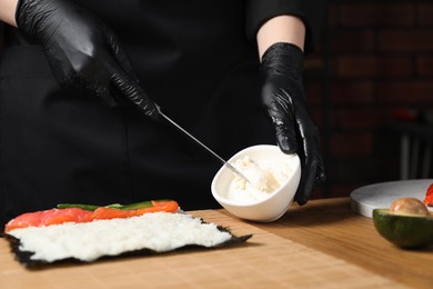 Photo of Chef in gloves putting cream cheese onto unwrapped sushi roll at wooden table, closeup
