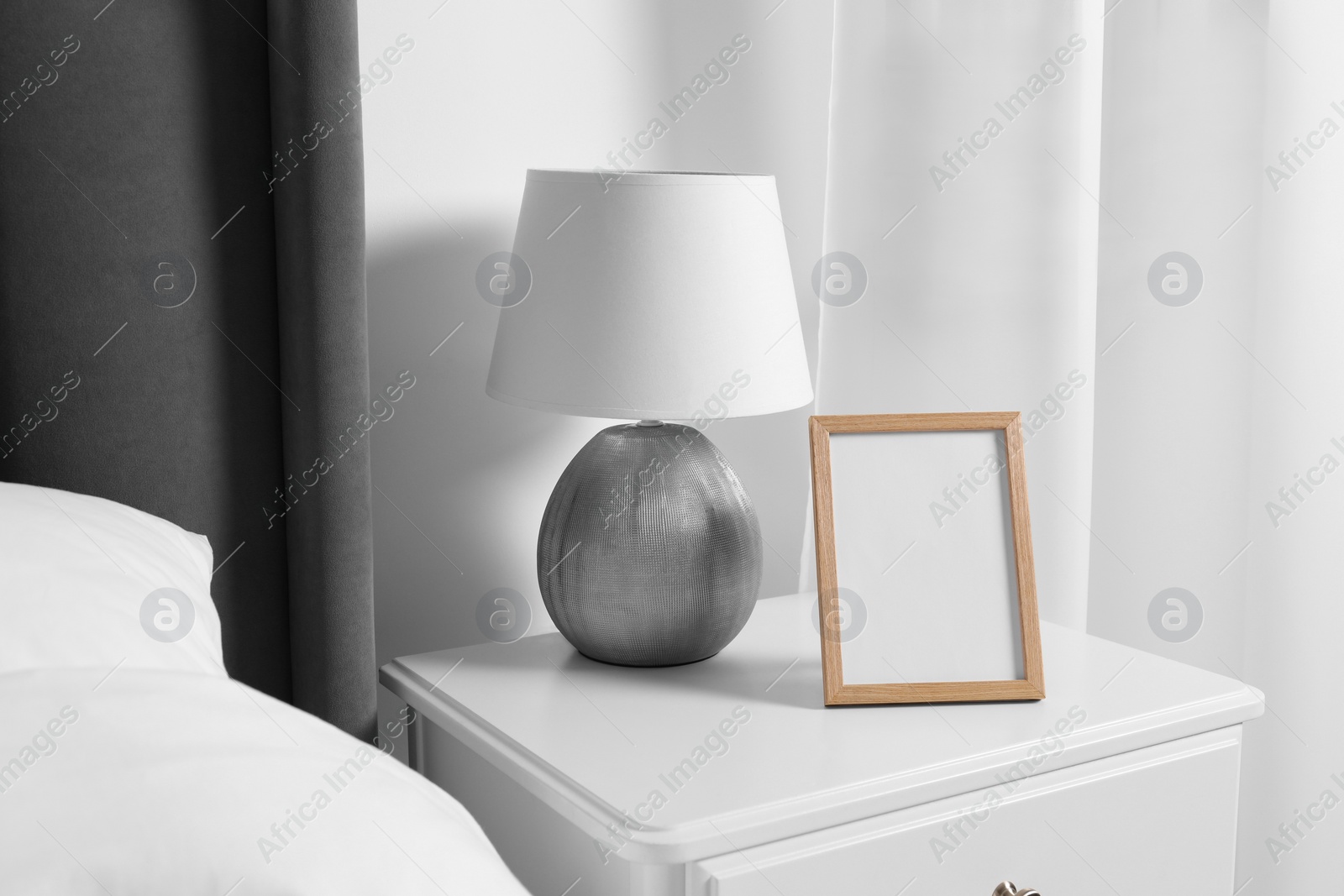 Photo of Empty square frame and lamp on white bedside table in room