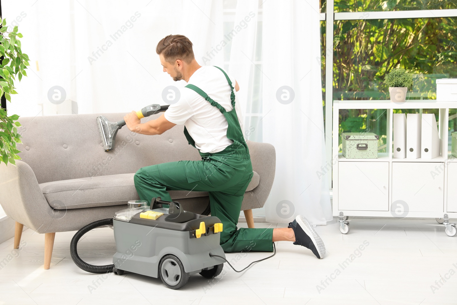 Photo of Male janitor removing dirt from sofa with upholstery cleaner in room