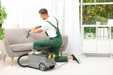 Male janitor removing dirt from sofa with upholstery cleaner in room