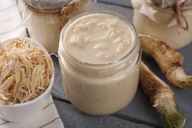 Photo of Spicy horseradish sauce in jars and roots on grey wooden table, closeup