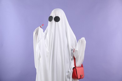Glamorous ghost. Woman in white sheet with stylish bag and lipstick on violet background