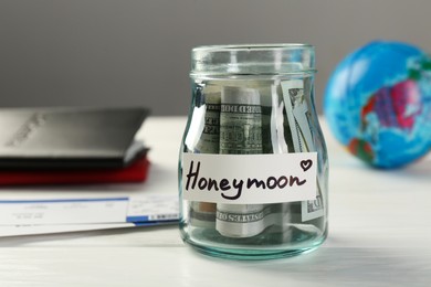 Glass jar with word Honeymoon and dollar banknotes on white wooden table, closeup