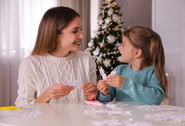 Happy mother and daughter making paper snowflakes at table near Christmas tree indoors