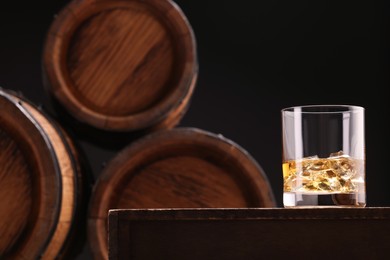 Photo of Whiskey with ice cubes in glass on wooden table near barrels against black background, space for text