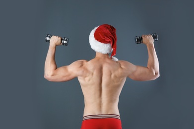 Photo of Young muscular man in Santa hat with dumbbells on gray background