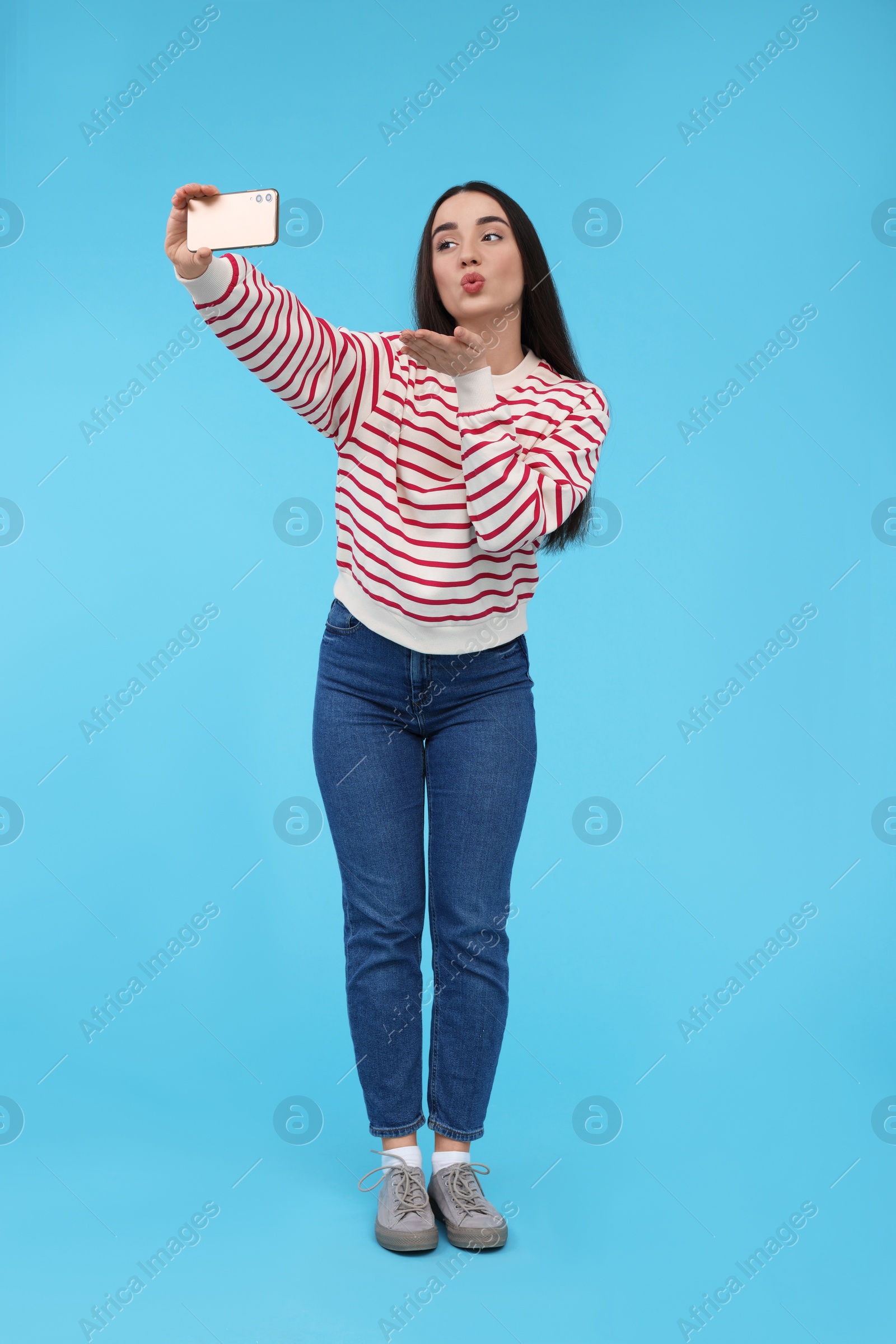 Photo of Young woman taking selfie with smartphone and blowing kiss on light blue background