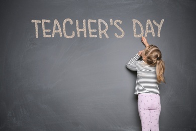 Cute little girl writing phrase Teacher's Day with chalk on blackboard