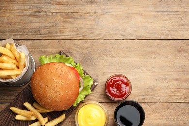 French fries, tasty burger, sauces and drink on wooden table, flat lay. Space for text