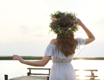 Photo of Young woman wearing wreath made of beautiful flowers on pier near river, back view