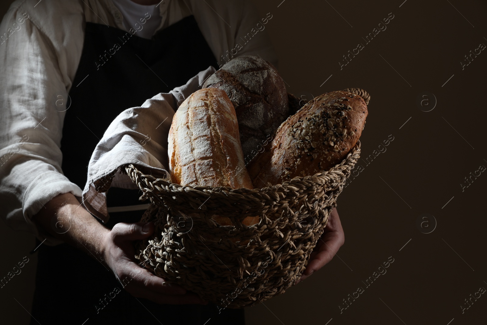 Photo of Man holding wicker basket with different types of bread on dark brown background, closeup