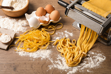 Photo of Pasta maker machine with dough and products on wooden table, above view