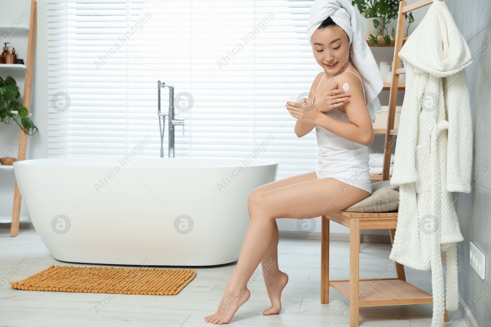 Photo of Beautiful young Asian woman applying body cream on shoulder in bathroom, space for text