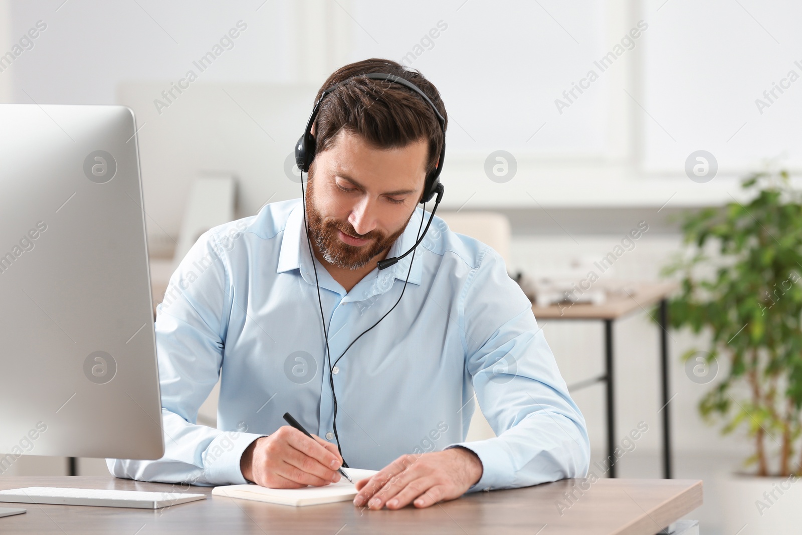 Photo of Hotline operator with headset writing something in notebook while working at wooden table