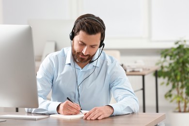 Hotline operator with headset writing something in notebook while working at wooden table