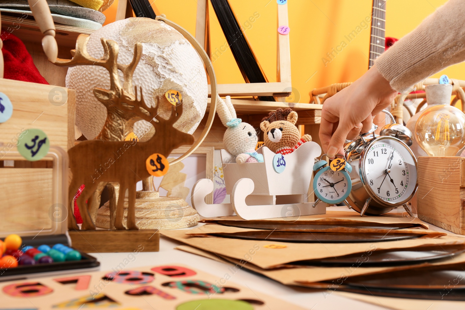 Photo of Woman holding alarm clock near table with many different stuff indoors, closeup. Garage sale