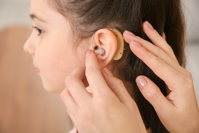 Photo of Young woman putting hearing aid in daughter's ear indoors, closeup