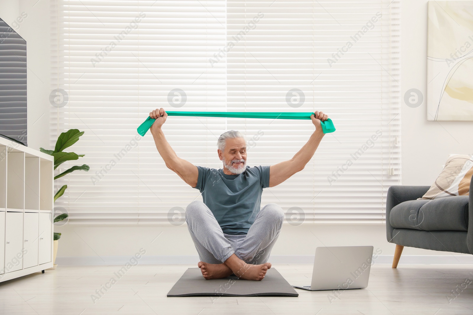 Photo of Senior man doing exercise with fitness elastic band near laptop on mat at home