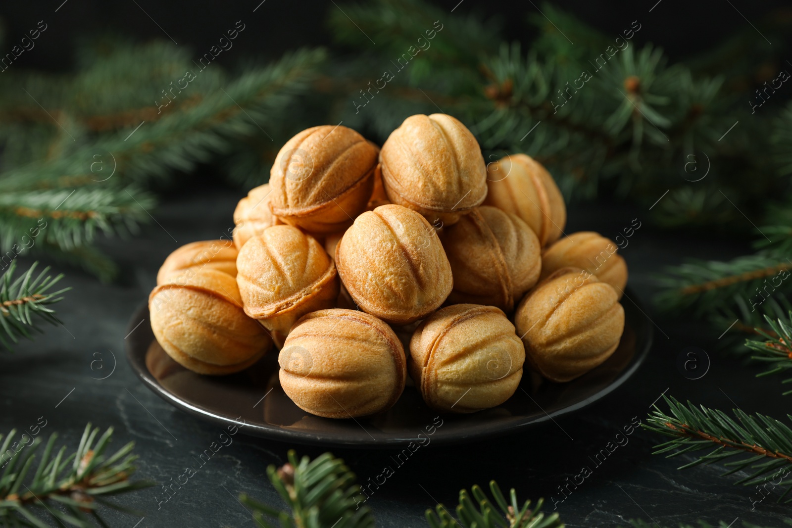 Photo of Plate of tasty nut shaped cookies among fir branches on black table, closeup