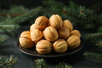 Plate of tasty nut shaped cookies among fir branches on black table, closeup